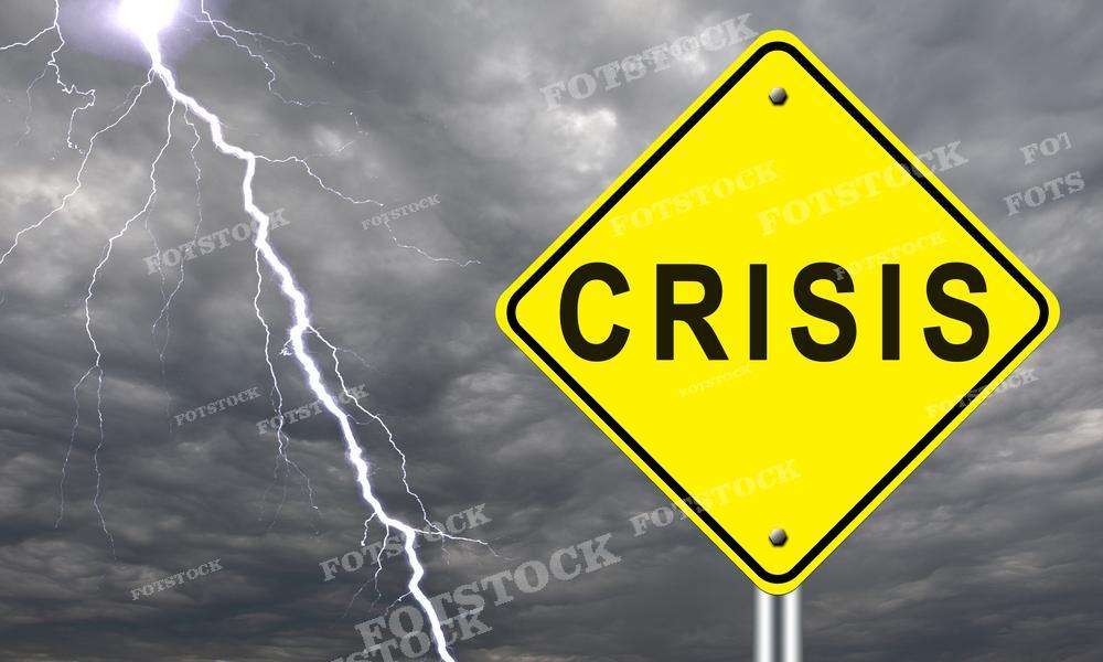 На фотографии изображён предупреждающий знак жёлтого цвета с надписью "CRISIS", который находится на переднем плане. В фоне небо затянуто тучами, и сквозь них видны молнии, подчёркивающие серьёзность и напряжённость ситуации, символизируемой знаком. Такое изображение может быть использовано для иллюстрации статей о финансовом кризисе, экономических трудностях, управлении рисками или в новостных материалах, посвящённых кризисным явлениям в экономике и политике.